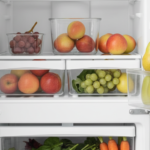 Pessoa organizando morangos, folhas verdes, cenouras e outras frutas e verduras em uma geladeira doméstica limpa e clara.