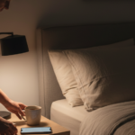 Pessoa organizando um livro e uma xícara no criado-mudo de um quarto com luz baixa, preparando uma rotina de sono tranquila.